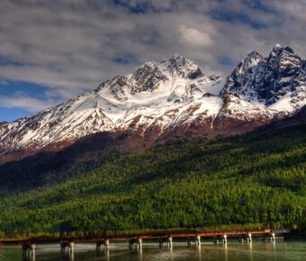 Snowy mountain peaks, lush green valley, tranquil river, wooden bridge, serene landscape.
