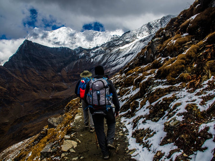 Hikers on narrow alpine trail with snow-capped mountains in the background.