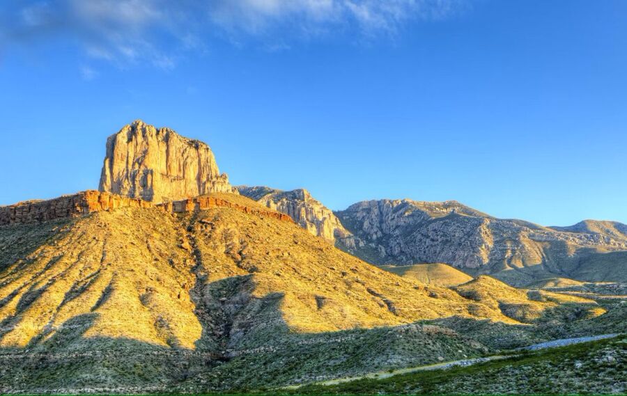 Sunlit Guadalupe Mountains landscape with rugged butte, desert plants, and clear blue sky.