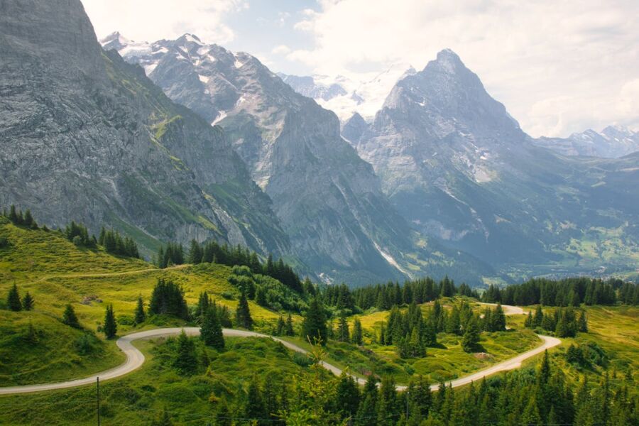 Alpine landscape with snow-capped peaks, lush greenery, winding road, and inviting blue sky.