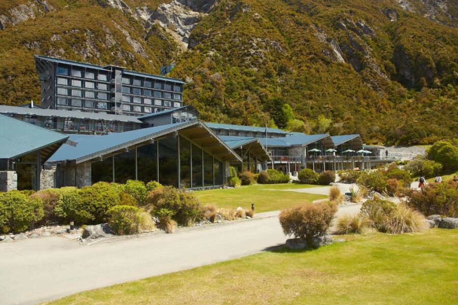Luxury mountain lodge resort with modern hotel buildings, green lawns, dining terrace, and dramatic alpine backdrop.