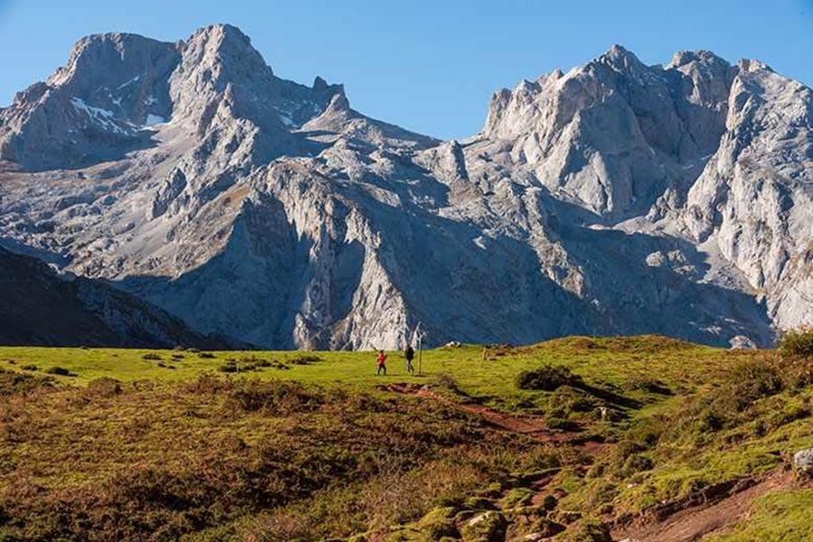 Panoramic Picos de Europa mountains with hikers crossing a grassy meadow beneath sheer rock faces.