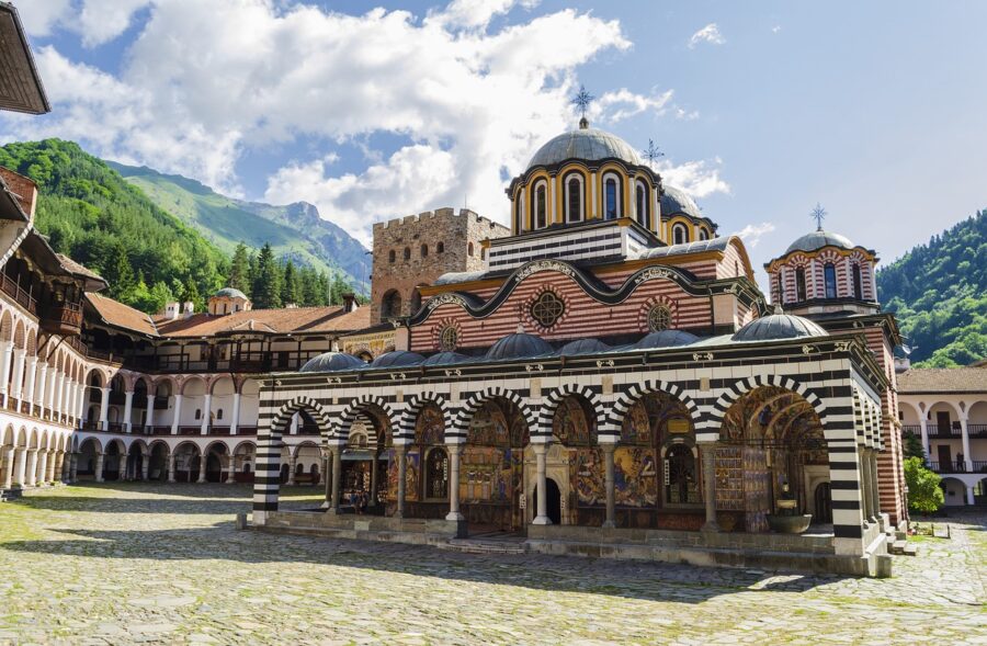 Rila Monastery courtyard in Bulgaria with domed church, striped arches, frescoes, and mountain backdrop.