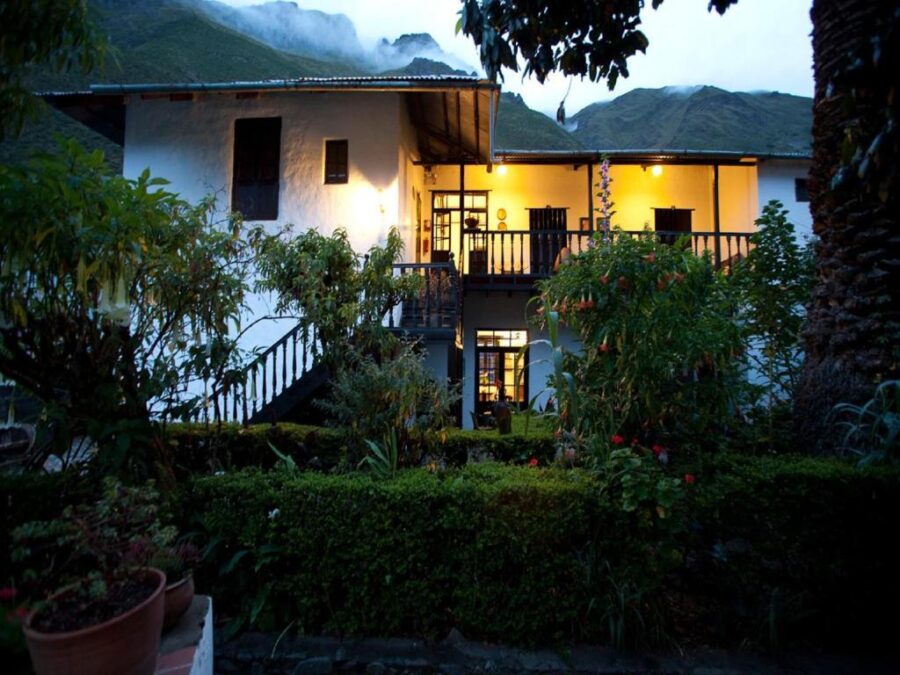 Rustic mountain inn at dusk with warm lights, balcony, lush garden, and misty green peaks.