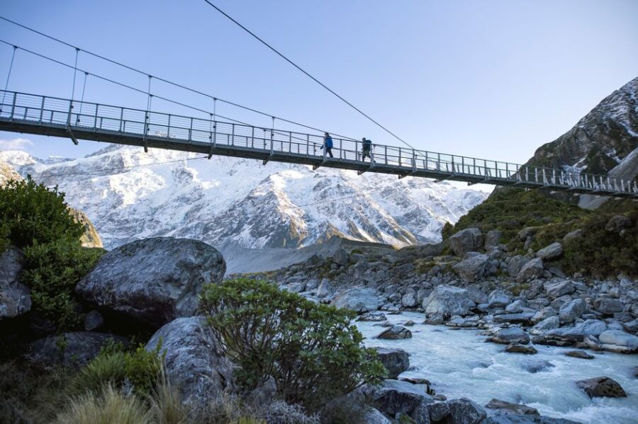 Hikers crossing mountain suspension bridge over glacial river with snowy alpine peaks in Hooker Valley.