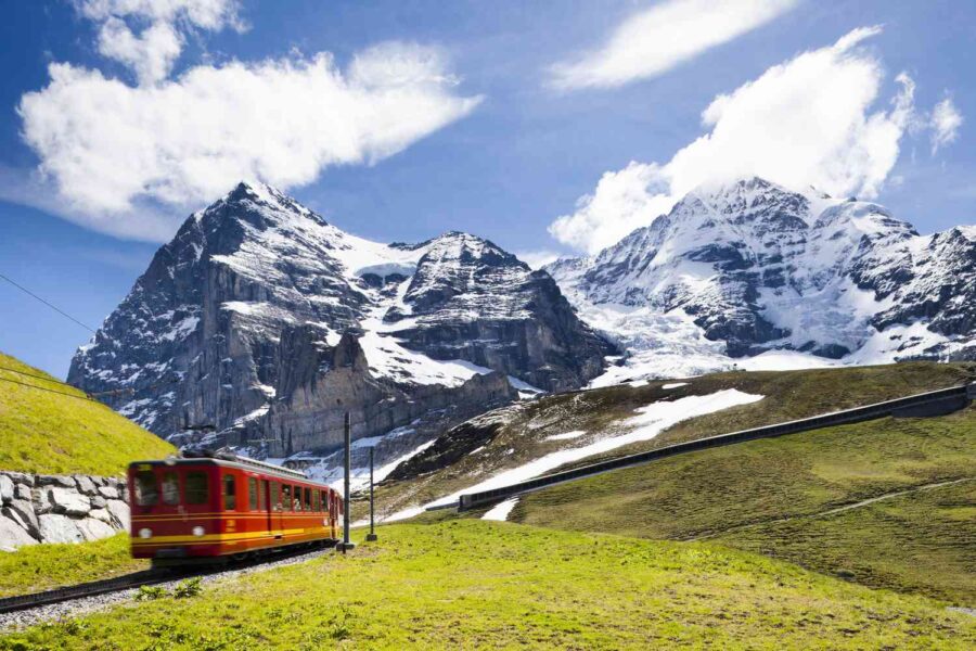 Red train winds through lush mountains under sunlit, snow-capped peaks and blue sky.