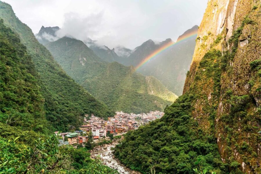 Scenic valley town amidst lush mountains with a rainbow and river.