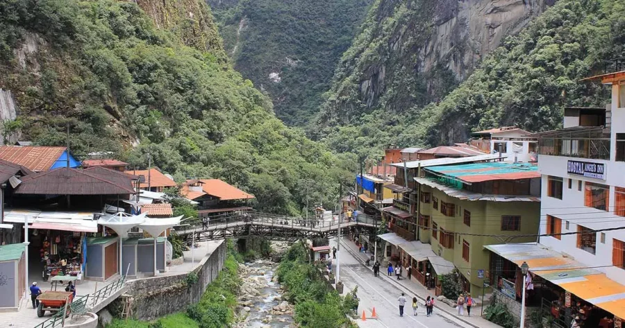 Aguas Calientes mountain town with river, wooden bridge, steep green Andes valley and tourist hotels.