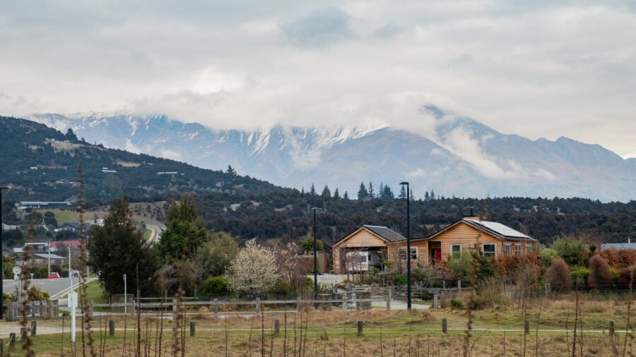 Mountain village landscape with wooden houses, rural field, winding road and dramatic snow-capped mountains.