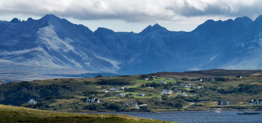 Coastal village with white houses, lush greenery, and majestic mountain backdrop under a cloudy sky.
