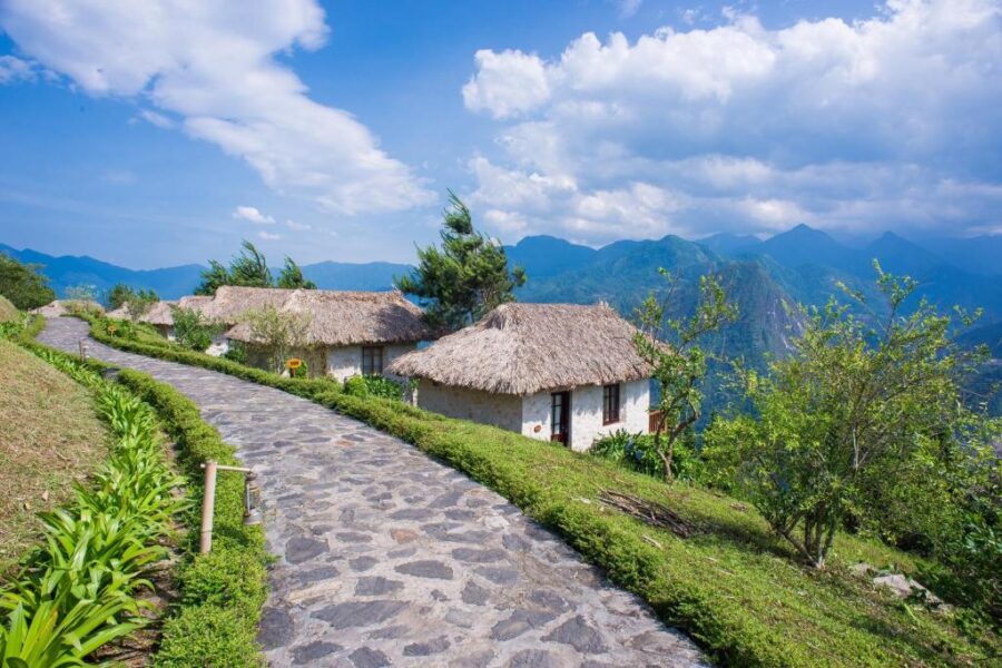 Mountain ecolodge village with stone pathway, thatched cottages, and scenic forested mountains at sunrise.
