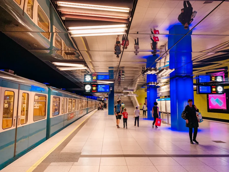 Munich U-Bahn platform with teal train, mirrored ceiling, blue-lit columns, and passengers.