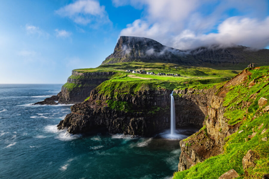 Mulafossur Waterfall cascading down cliffs into a lush green valley in the Faroe Islands, surrounded by dramatic landscapes.
