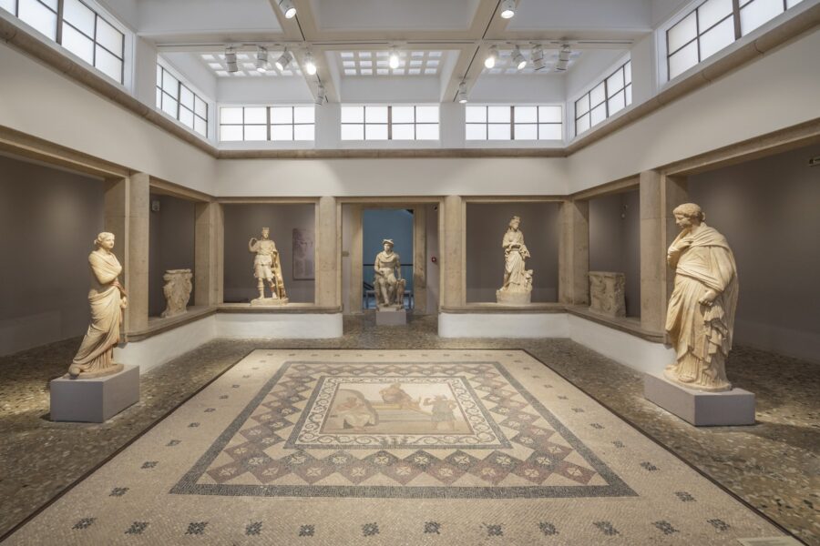 Museum gallery with mosaic floor and classical marble statues under skylights.