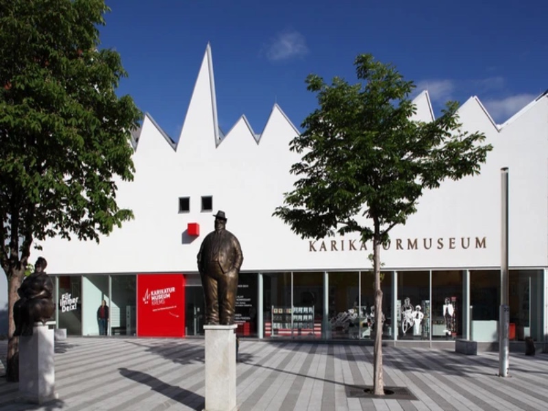 Karikaturmuseum modern museum with white façade, sculptures, and plaza under blue sky.