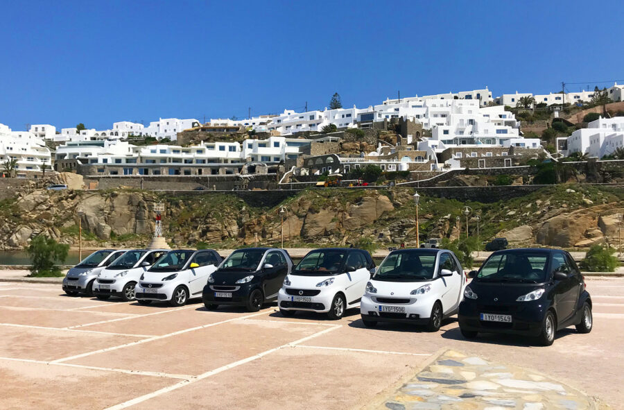 Mykonos car rental with small cars and SUVs against iconic white-washed buildings backdrop.