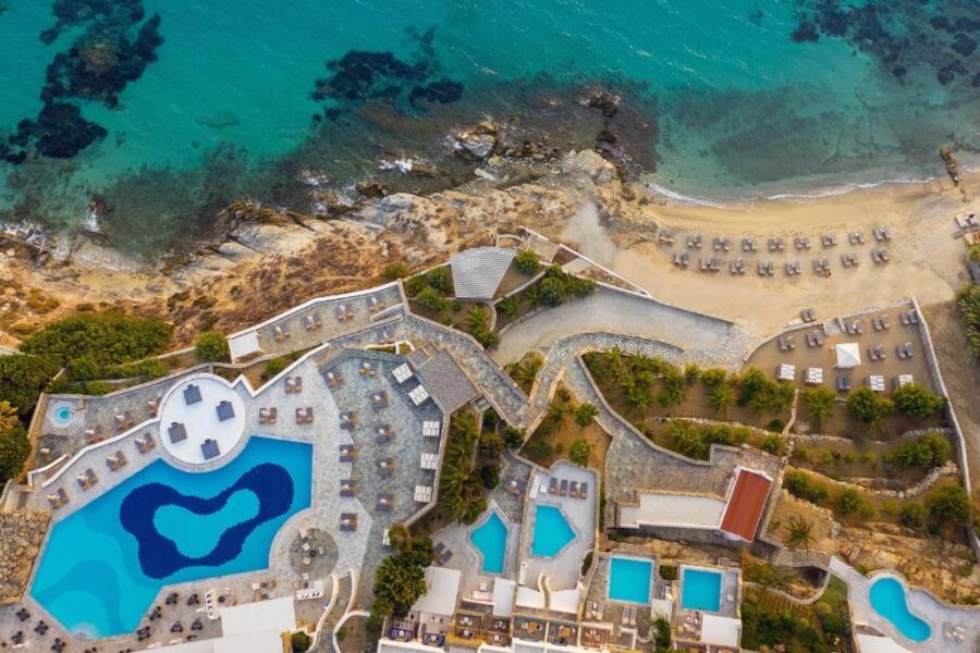Aerial view of the beach and pool at Mykonos Grand Hotel & Resort, showcasing the vibrant blue water and sandy shore.
