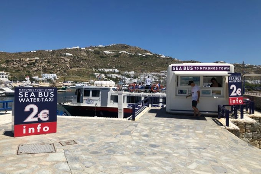 Mykonos waterfront with Sea Bus water-taxi, cobblestone walkway, and hillside white buildings.