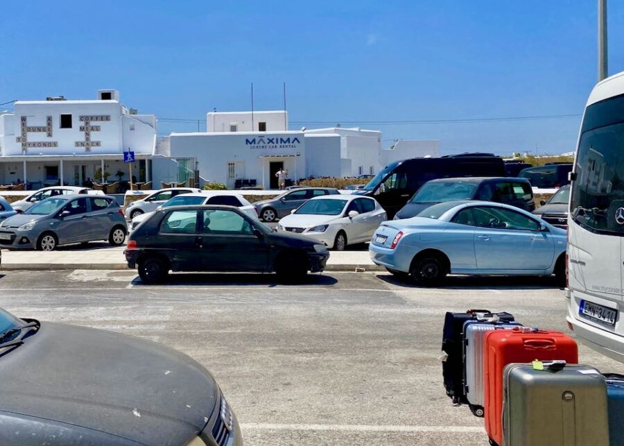 Vibrant Mykonos parking scene with travelers, luggage, rental cars, and coastal architecture.