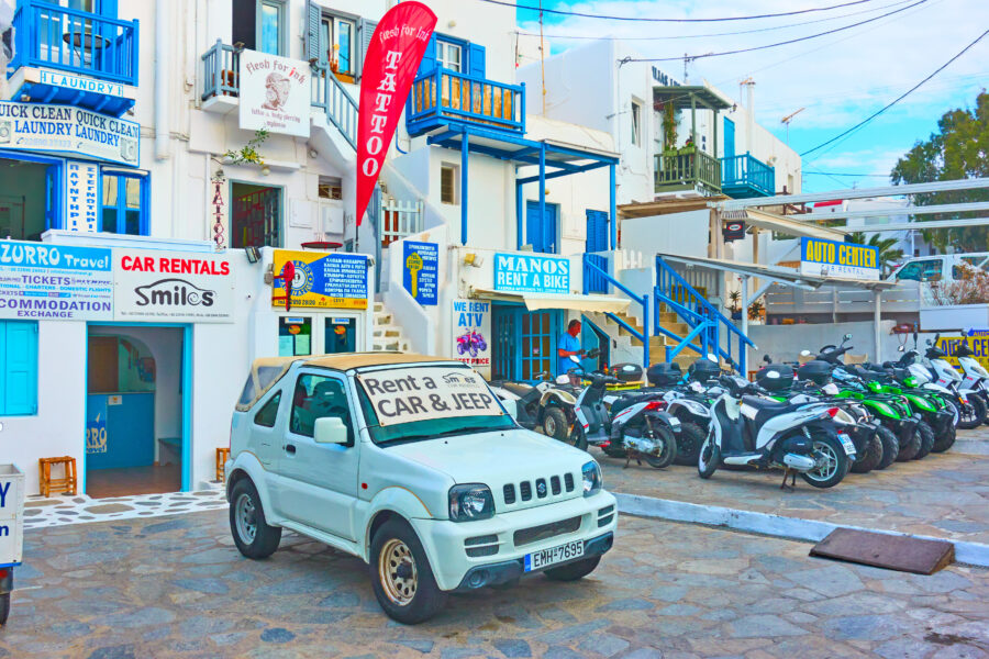 Scenic view of Mykonos town, Greece, featuring a car rental service with vibrant buildings and the Aegean Sea in the background