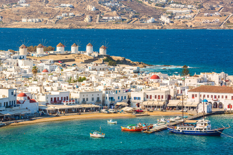 Mykonos' Chora port featuring a red church, iconic windmills, and yachts on a bright summer day by the Aegean Sea