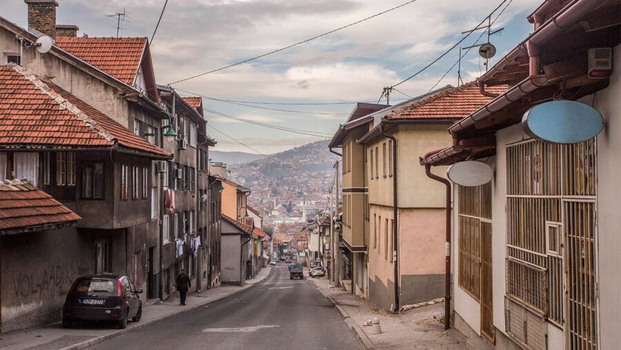 Steep narrow Sarajevo street with old hillside houses, parked cars, and misty valley city view.