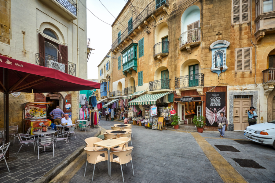 Victoria, Gozo narrow stone street with outdoor cafes, market stalls, colorful awnings and historic balconies.