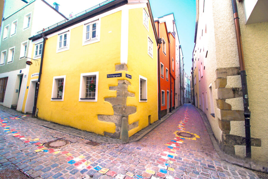 Sunlit narrow cobbled alley in Passau with yellow corner house and painted pathway.