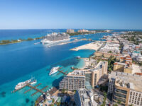 Aerial view of a Nassau bustling port city with a large cruise ship docked in turquoise waters. Skyscrapers line the coast under a bright blue sky, conveying a lively, tropical atmosphere.