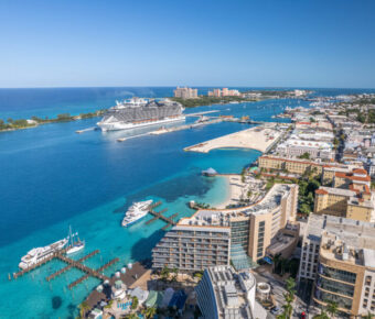 Aerial view of a Nassau bustling port city with a large cruise ship docked in turquoise waters. Skyscrapers line the coast under a bright blue sky, conveying a lively, tropical atmosphere.