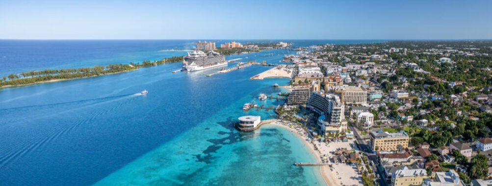 Aerial view of a vibrant Nassau coastal city with a cruise ship docked in azure waters. Sandy beaches lined with resorts on a sunny day evoke a lively, tropical atmosphere.