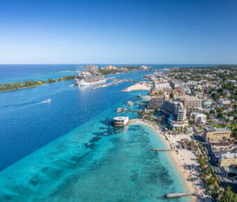 Aerial view of a vibrant Nassau coastal city with a cruise ship docked in azure waters. Sandy beaches lined with resorts on a sunny day evoke a lively, tropical atmosphere.