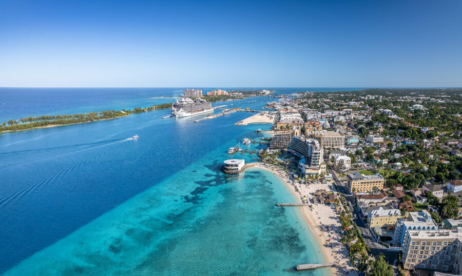 Aerial view of a vibrant Nassau coastal city with a cruise ship docked in azure waters. Sandy beaches lined with resorts on a sunny day evoke a lively, tropical atmosphere.