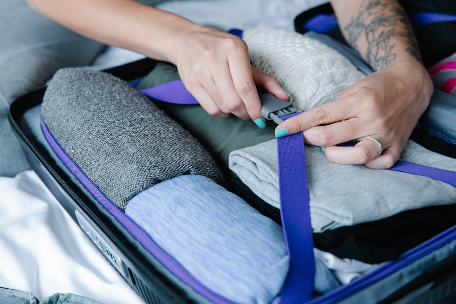 Woman tightening compression strap on neatly packed travel suitcase with folded clothes and cozy sweater.