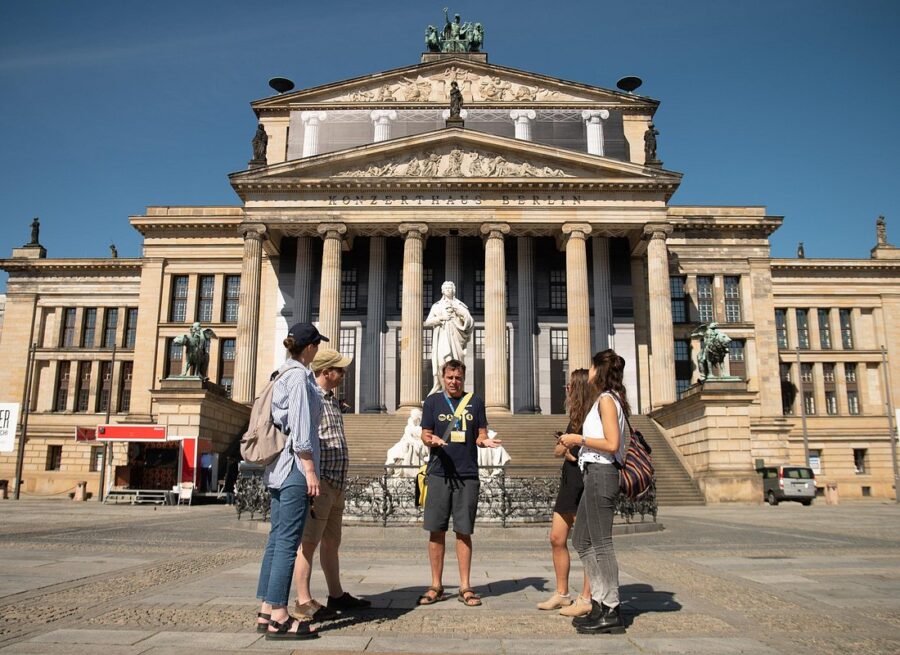 Berlin neoclassical building with columns, statue on steps, tourists in sunlit square.