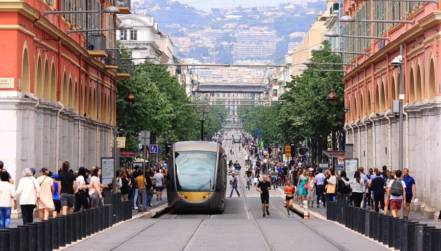 Modern tram in vibrant city street with diverse pedestrians and elegant architecture in Nice, France.