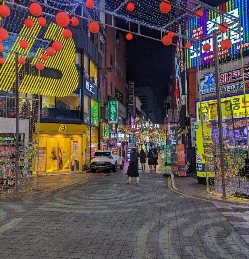 Vibrant Korean nightlife street with neon signs, food alley restaurants, and illuminated shopping district at night.