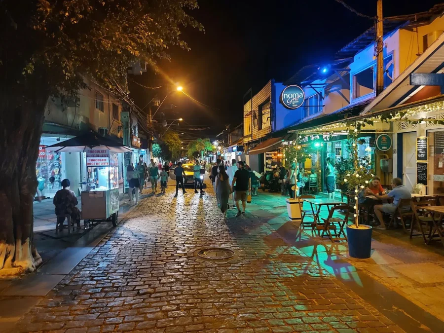 Night cobblestone street with outdoor restaurants, people dining, street food cart, and colorful lights.