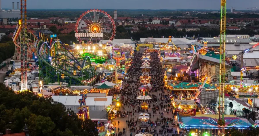 Oktoberfest nighttime aerial view of crowded fairground with Ferris wheel and roller coaster.