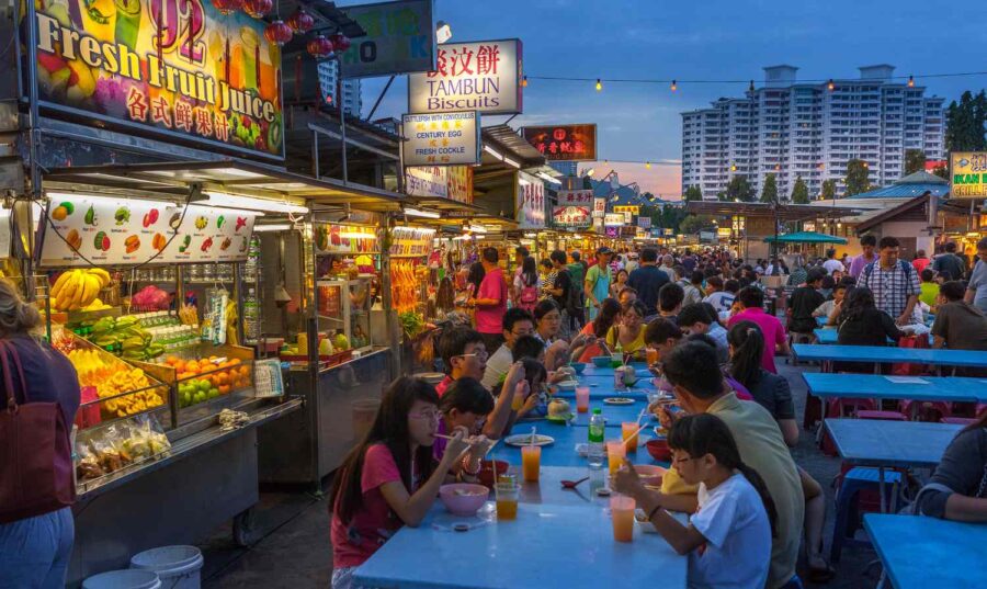 Crowded Penang night market with street food stalls, plastic tables, families dining under hanging lights.