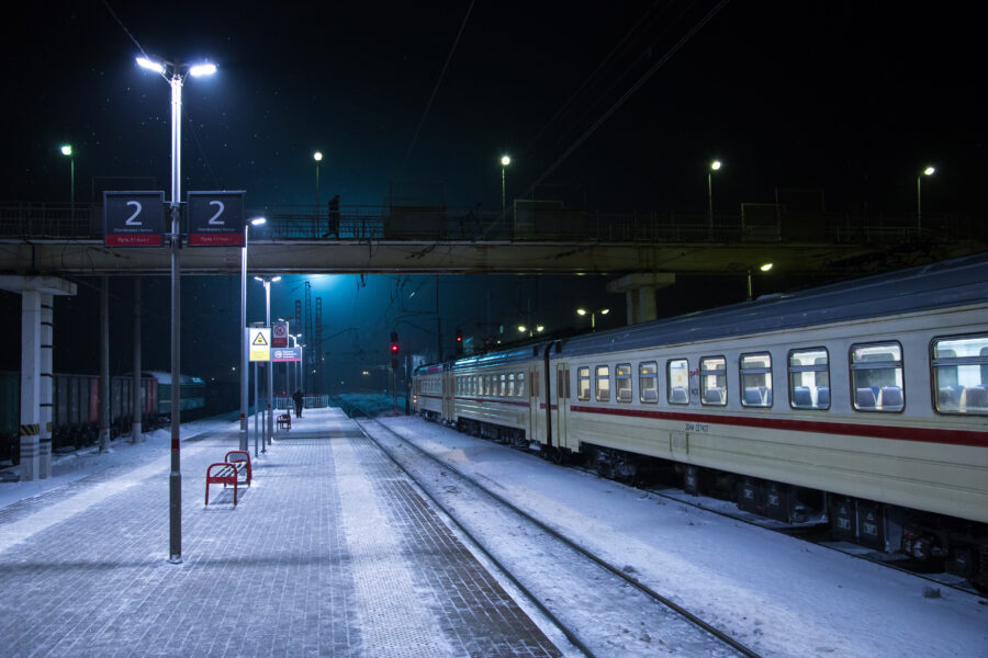 Winter night scene of a railway platform with a train illuminated by station lights. Snow covers the ground.