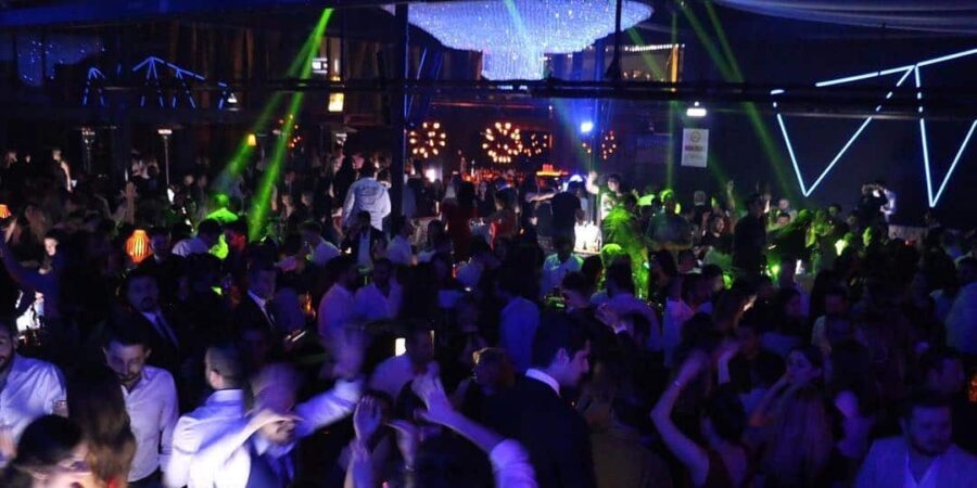 Crowded nightclub dance floor with neon lights, chandelier, and dancing people.