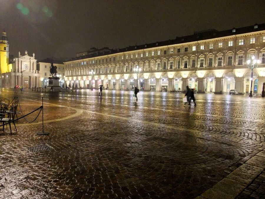 Rainy nighttime urban square with historic architecture, clock tower, and statue in Europe.