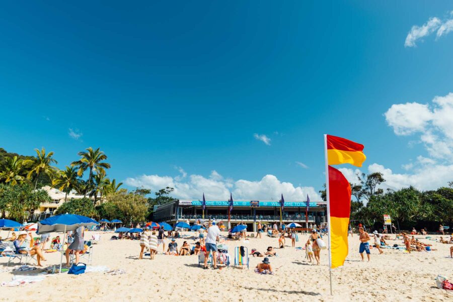 Sunny Noosa beach with red yellow surf lifesaving flag, umbrellas, crowds, blue sky.