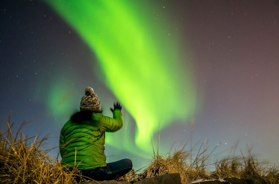 Individual seated on the ground, arms lifted, admiring the stunning aurora borealis illuminating Reykjavik's night sky.