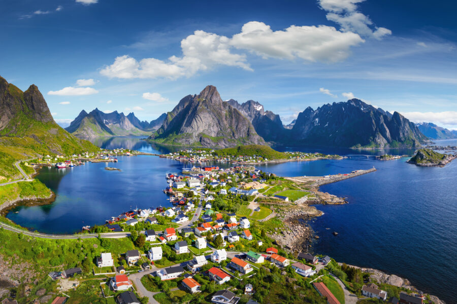 Scenic view of Reine village in Lofoten, Norway, featuring traditional rorbu houses against a sunny, blue sky backdrop.