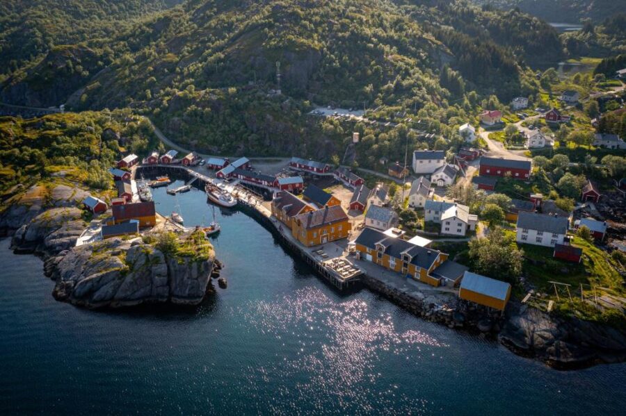Aerial view of a Nusfjord Village & Resort with colorful houses near a small harbor. Green hills surround the area under a clear blue sky.
