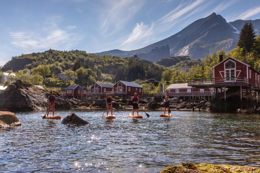 People paddle board on calm water in front of a mountain near Nusfjord Village & Resort.