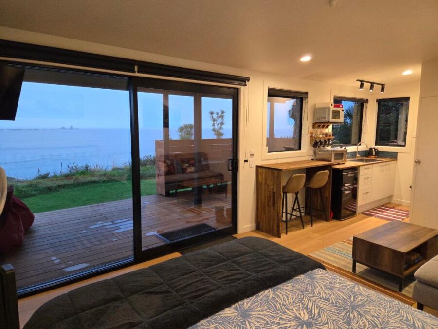 Modern oceanfront studio interior with bed, kitchenette, and panoramic sea view through large glass doors.