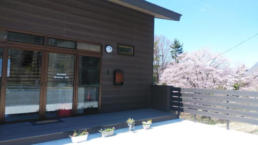 Okunikko guesthouse exterior with dark wood facade, glass entrance, deck, planters, and spring cherry blossoms.